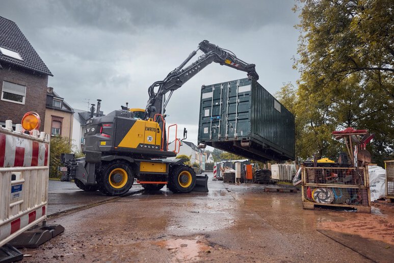 Pelle sur pneus compacte Volvo soulevant un conteneur, illustrant sa productivité élevée et sa maniabilité dans les environnements d'infrastructures étroits